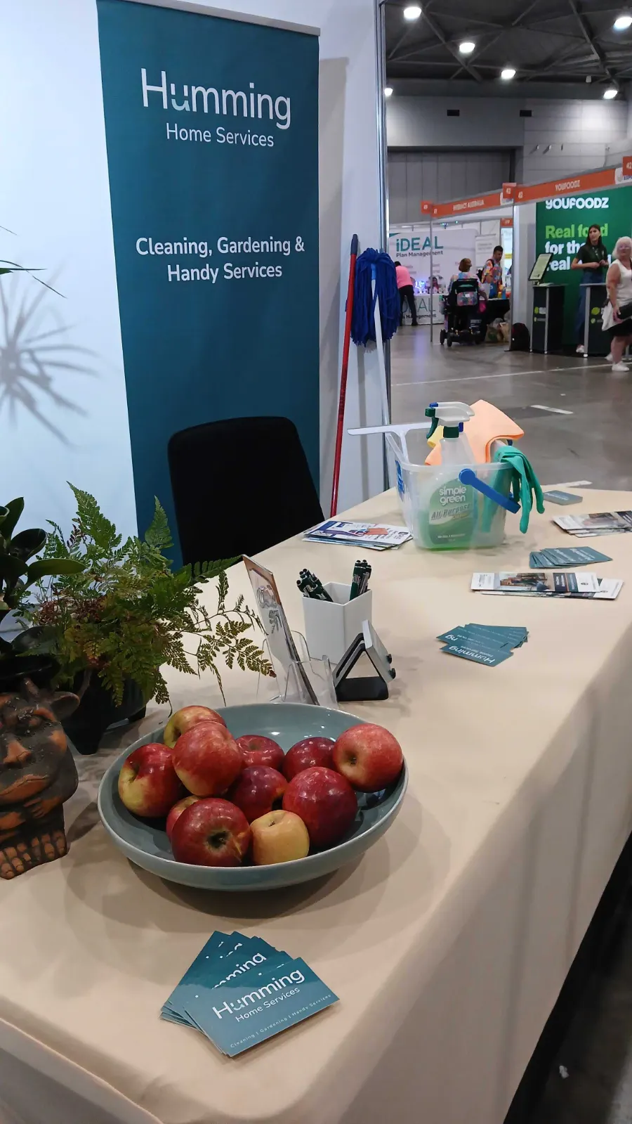 Humming Home Services expo table with business cards, brochures and cleaning caddy in front of a Cleaning, Gardening and Handy Services banner