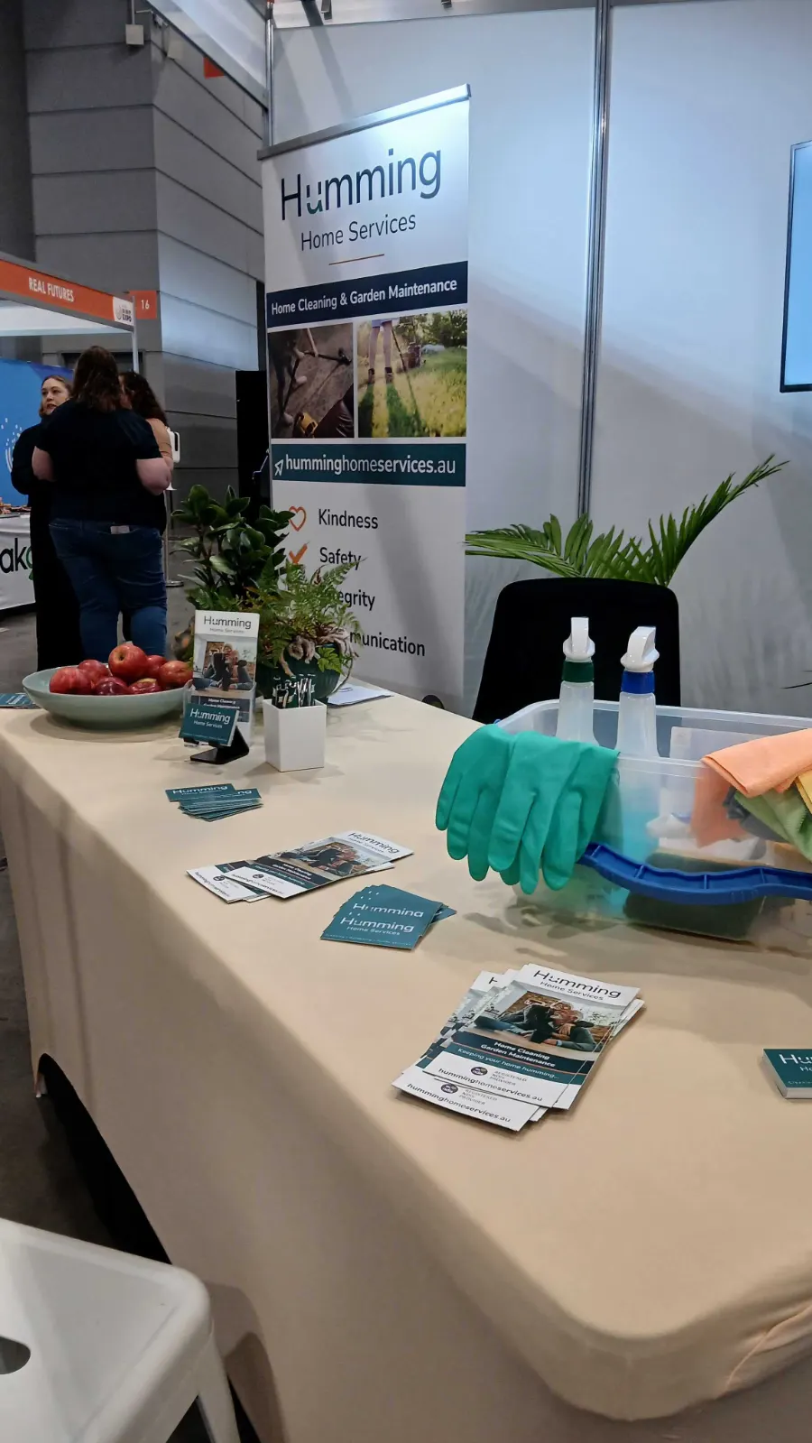 Humming Home Services brochures and business cards for home cleaning and garden maintenance on the expo table, with a banner listing kindness, safety, integrity and communication