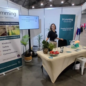 Humming Home Services team member smiling at the company booth at the Brisbane Disability Expo 2026, with banners showing its values and NDIS registered provider status