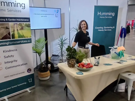 Humming Home Services team member smiling at the company booth at the Brisbane Disability Expo 2026, with banners showing its values and NDIS registered provider status