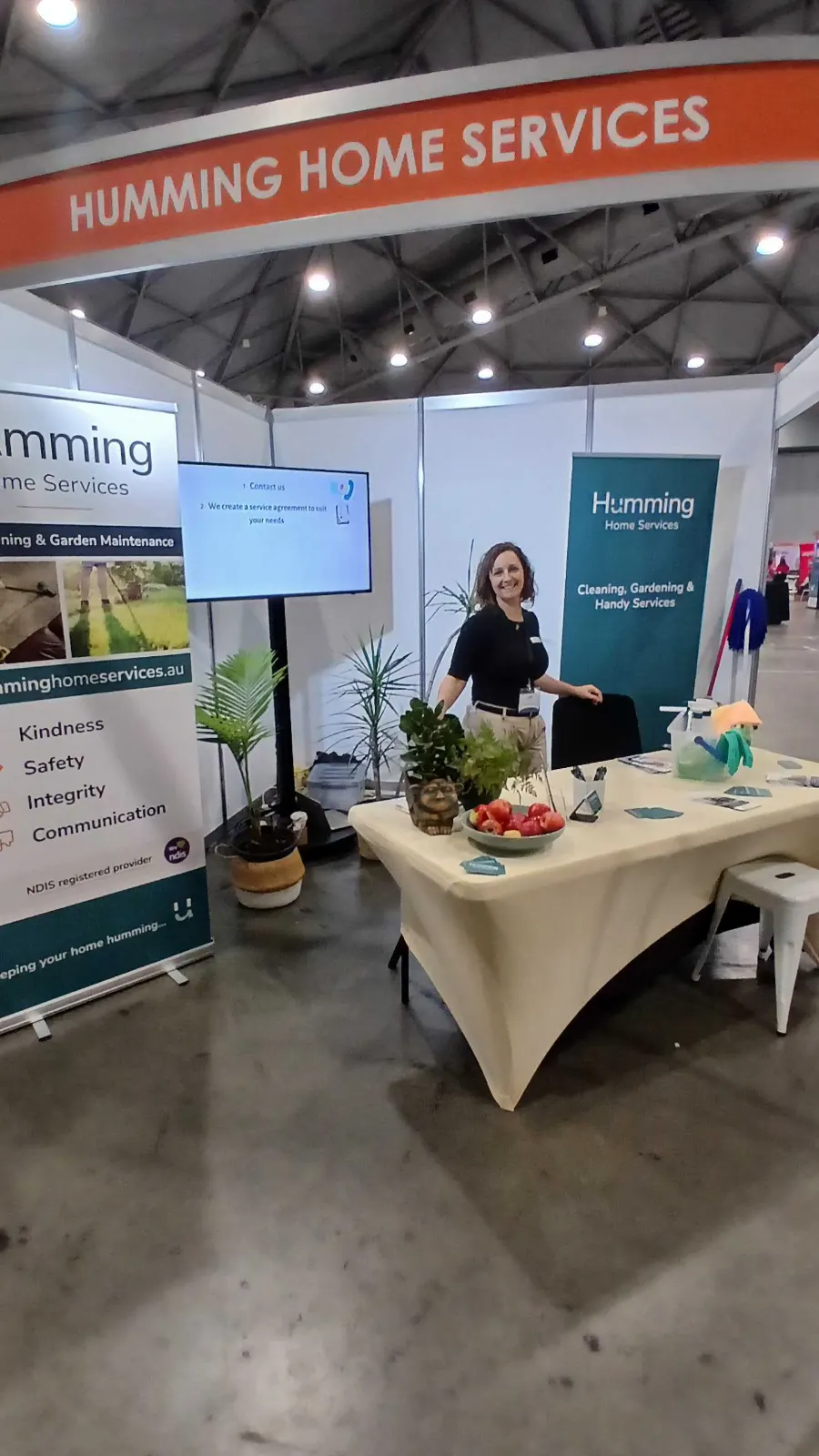 Humming Home Services team member smiling at the company booth at the Brisbane Disability Expo 2026, with banners showing its values and NDIS registered provider status